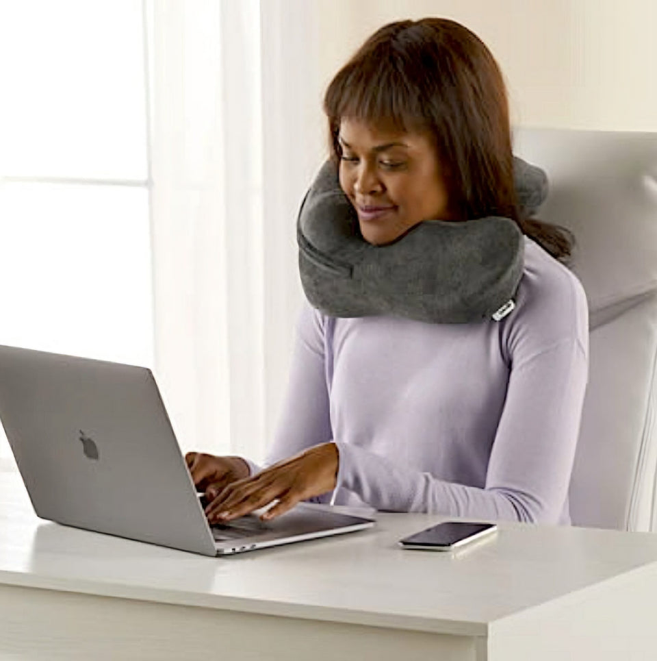 Woman using her grey Neck Sofa® Pillow while working on her computer at a white desk